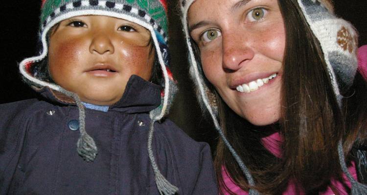 Close-up selfie van een reiziger en een lokaal kind die warm glimlachen in wollen oorflappenmutsen.