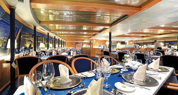 Helder, netjes ingedeeld restaurant aan boord van een schip met blauwe vloerbedekking en tafels met witte servetten.