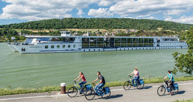 Fietsers rijden parallel aan een passerende riviercruiseschip met glooiende groene heuvels op de achtergrond.