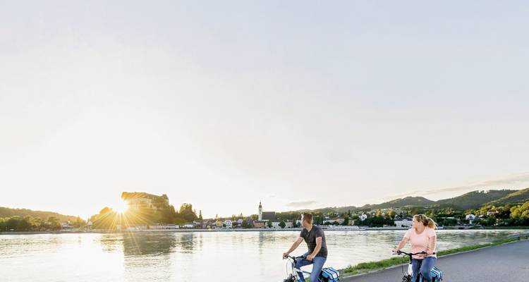 Fietsers rijden langs een rivieroever bij zonsondergang met zonnestralen over een verre kasteelheuvel.