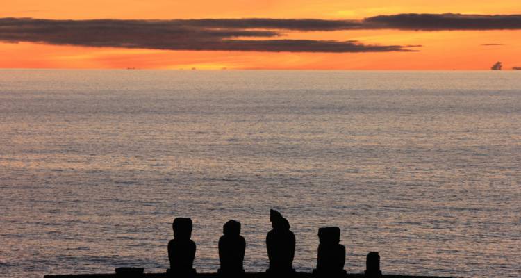 Vista del atardecer sobre el océano con estatuas Moai silueteadas contra el cielo.