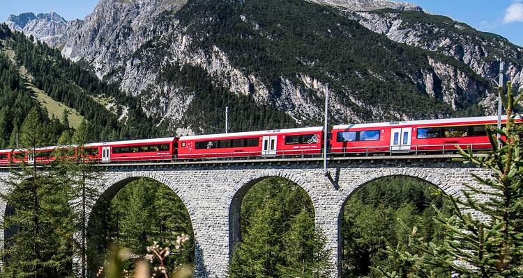 Tren rojo cruzando un viaducto de piedra alto con bosque denso y montañas al fondo.