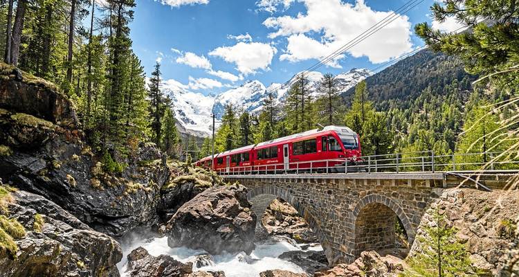Train rouge traversant un pont de pierre au-dessus d'une rivière tumultueuse, entouré d'une forêt luxuriante avec des pics enneigés au loin.