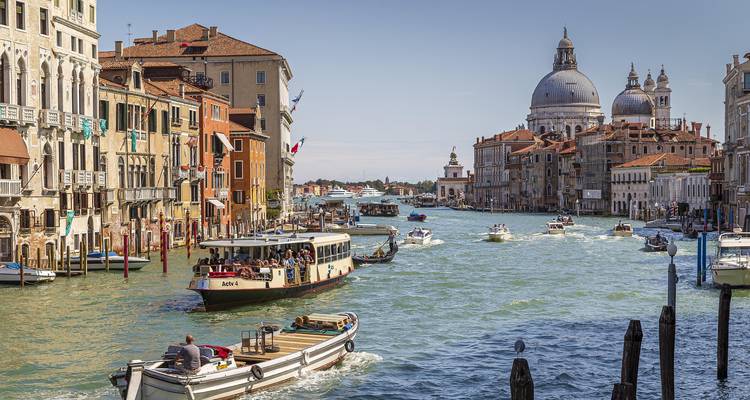 Canal veneciano animado con embarcaciones y edificios bulliciosos a ambos lados bajo un cielo brillante.