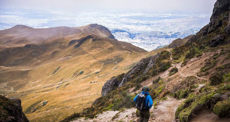 Hiker in blue jacket descending a rugged Andean trail with sweeping views over valleys and the city far below