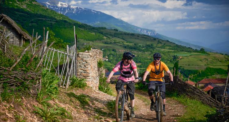 Dos ciclistas de montaña recorren un sendero agreste del campo con colinas verdosas y picos nevados detrás.