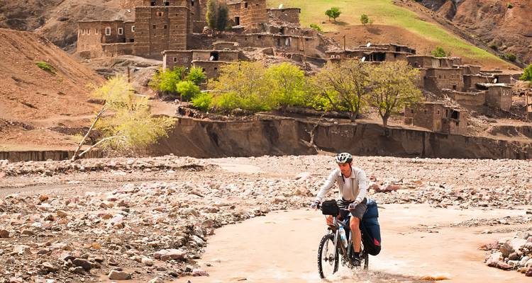 Ciclista vadea un río poco profundo y fangoso con un pueblo de montaña remoto al fondo.
