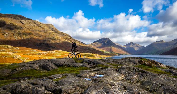 Un ciclista de montaña se encuentra triunfante sobre un afloramiento rocoso con vista a un lago y picos ondulantes bajo un cielo azul brillante.