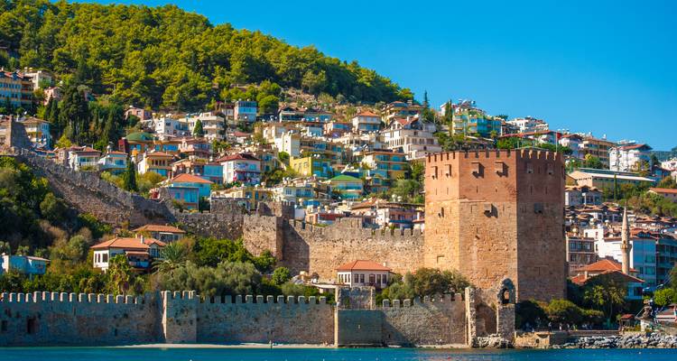 Antalyas Roter Turm und historische Seemauern vor einem Hügel mit bunten Häusern und grünen Pinien.