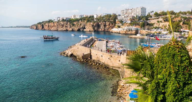 Klares türkisfarbenes Wasser und alte Hafenmauern von Antalya mit Booten und fernen Klippen.