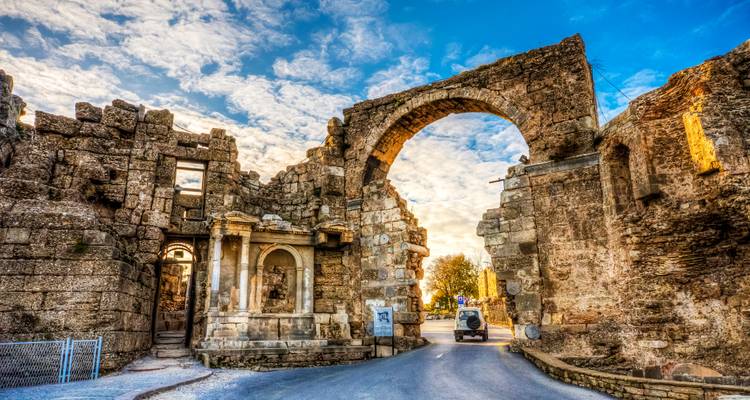 Antikes steinernes Torbögen von Side, Türkei, beleuchtet von warmem Abendlicht mit einem kleinen Fahrzeug, das hindurchfährt.