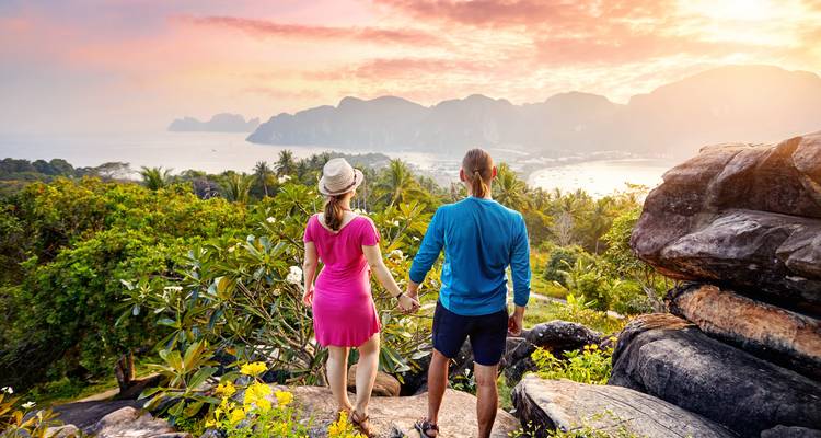 Junges Paar hält Händchen und genießt ein lebendiges Küstenbergpanorama bei Sonnenuntergang.