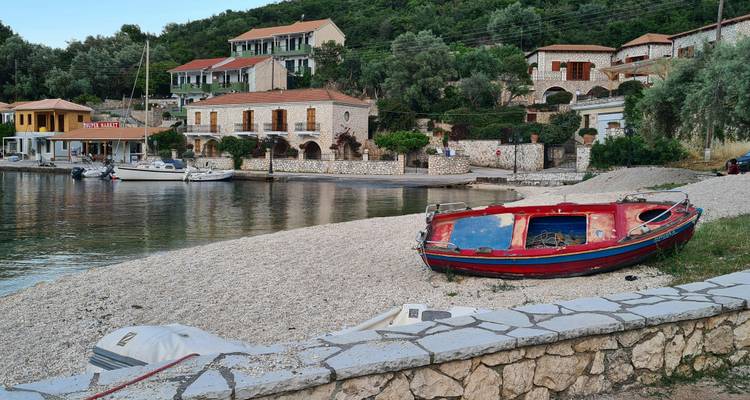 Tranquilo pueblo costero con casas de piedra en una ladera boscosa, una playa de guijarros y un bote de remos rojo varado en la orilla junto a aguas tranquilas.