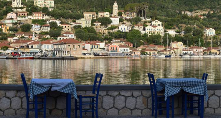 Mesas de taberna junto al agua y sillas azules dan a un puerto sereno bordeado de casas en tonos pastel y yates amarrados.