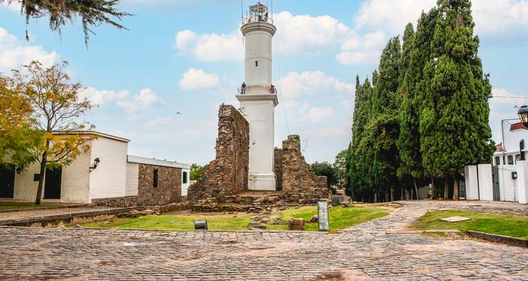 Phare avec ruines et chemin pavé, flanqué de grands arbres et de bâtiments historiques adjacents.