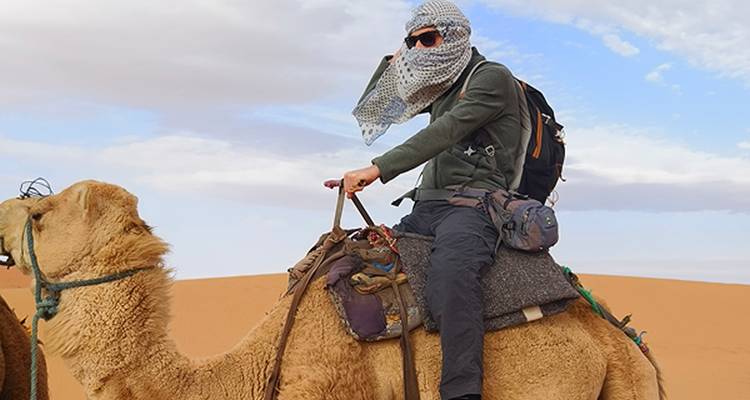 Reisender mit Schal und Sonnenbrille, der auf einem Kamel durch die Sanddünen der Sahara reitet