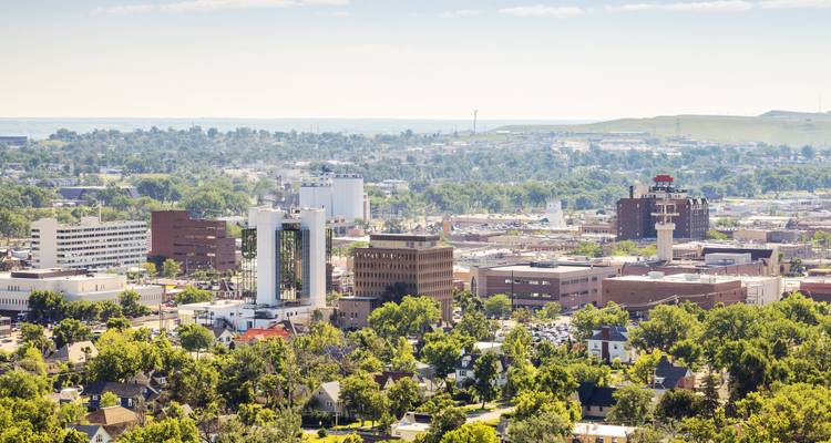 Aerial cityscape of Rapid City with modern buildings rising above leafy residential neighborhoods.