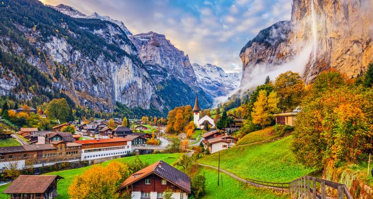 Malerisches Dorf mit Kirchturm liegt unter hoch aufragenden Klippen und Herbstwäldern in einem dramatischen Alpental.