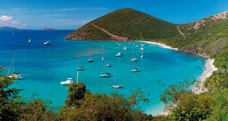 Baie turquoise avec de nombreux yachts ancrés près d'un promontoire boisé et d'une plage de sable blanc.