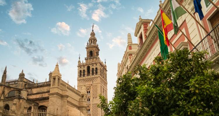 Blick nach oben auf den Glockenturm der Giralda und benachbarte historische Gebäude mit wehenden spanischen und andalusischen Flaggen vor einem blauen Abendhimmel.