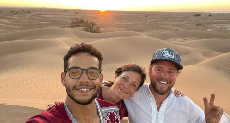 Smiling friends take a selfie on rolling sand dunes lit by a warm desert sunset.