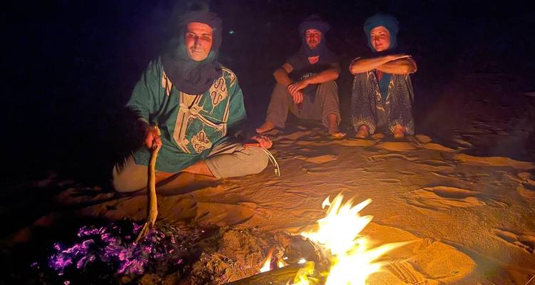 Berber guides sit by a small campfire at night, faces illuminated by the flames in the desert.