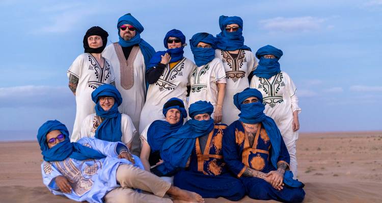 Tour group dressed in blue Tuareg robes poses on open desert sands under pastel evening light.