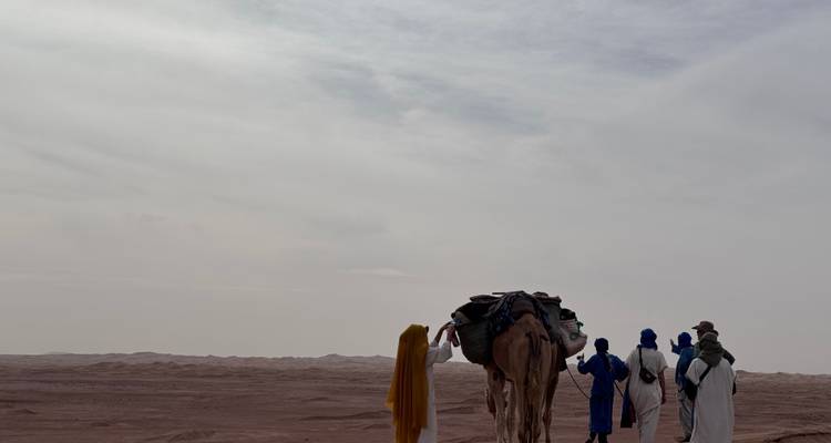 Camel caravan led by blue-robed guides charts a course across hazy desert flats.