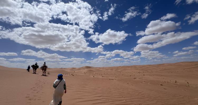 Travellers and camels walk across sweeping sand dunes beneath a sky dotted with puffy clouds.