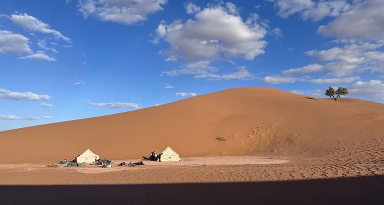 Two small tents and travellers rest at the foot of a towering orange sand dune under blue skies.