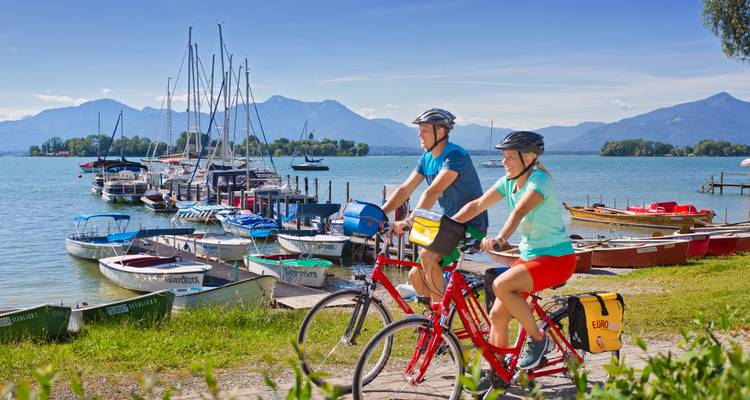 Cyclistes sur un sentier au bord d'un lac avec des bateaux et un paysage de montagne.