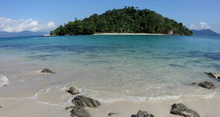 Klares, flaches Wasser, das gegen sandiges Ufer plätschert, mit einer kleinen bewaldeten Insel vor der Küste.
