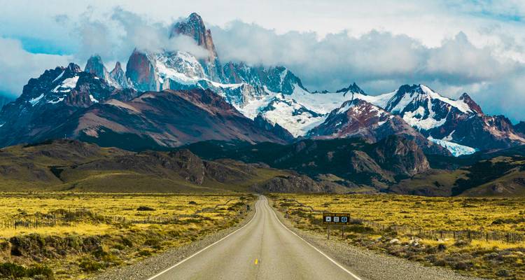 Une longue route menant vers des montagnes enneigées sous un ciel nuageux.