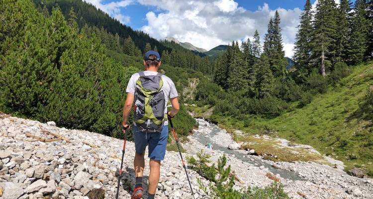 Randonneur marchant le long d'un sentier rocheux avec un ruisseau et des montagnes.