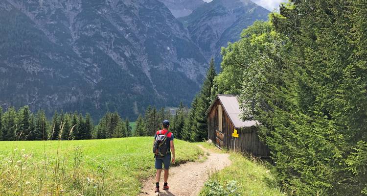 Randonneur sur un sentier près d'une cabane en bois avec des montagnes au loin.