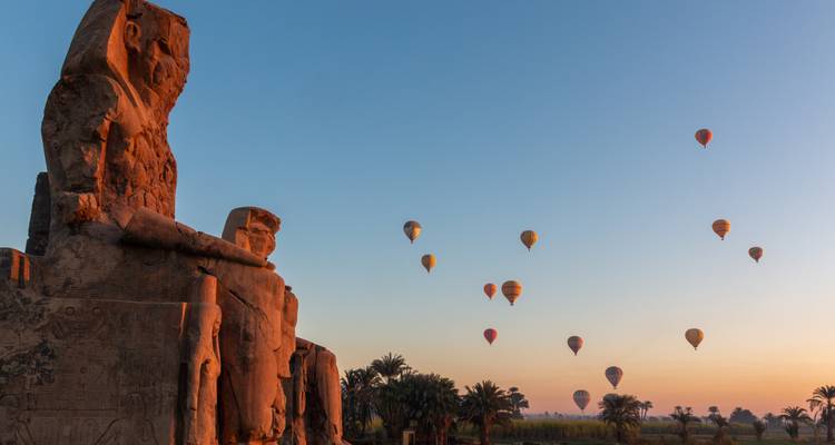 Kolosse von Memnon in Luxor als Silhouetten vor einem Sonnenaufgangshimmel mit bunten Heißluftballons.