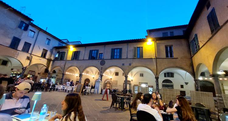 Escena nocturna de un patio italiano rodeado de arcos donde la gente cena en mesas al aire libre.