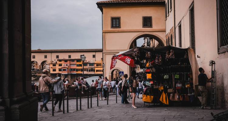 Puesto de mercado concurrido junto al río cerca del Ponte Vecchio con compradores y el famoso puente de fondo.