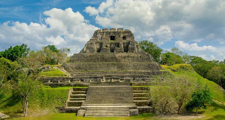 Ancienne pyramide maya à degrés s'élevant depuis des terrasses herbeuses sous des ciels bleus partiellement nuageux.