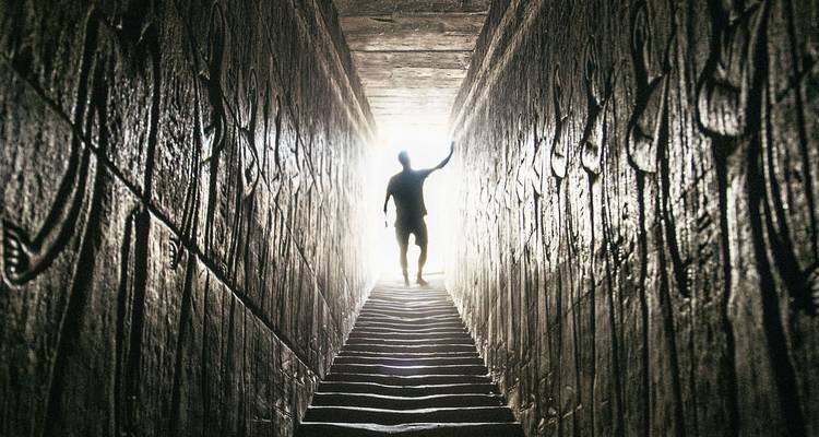 Silhouetted visitor climbs narrow ancient passageway toward bright daylight.