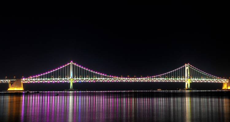 Vista nocturna de un puente iluminado con luces coloridas reflejándose en el agua.