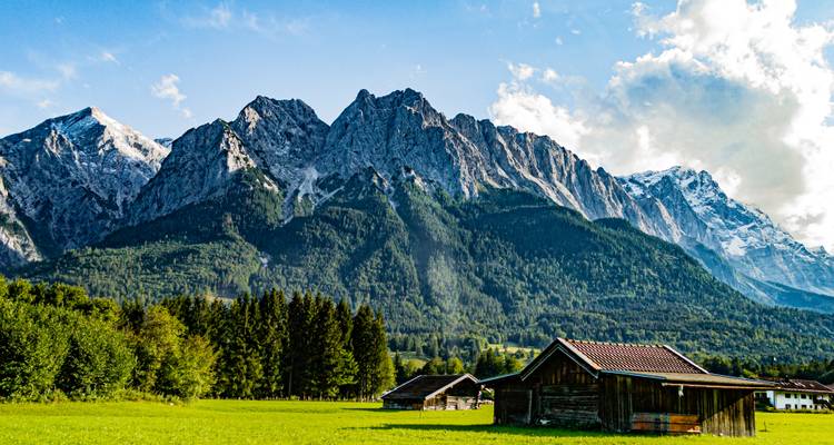 Hütten in einem grünen Feld mit sich erhebenden Bergen im Hintergrund unter einem blauen Himmel.