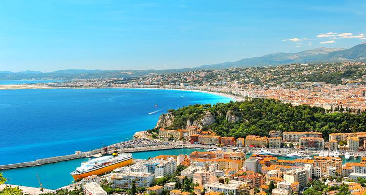 Panoramic aerial view of Nice with its sweeping azure bay, curving beachfront promenade and colorful cityscape backed by distant mountains.