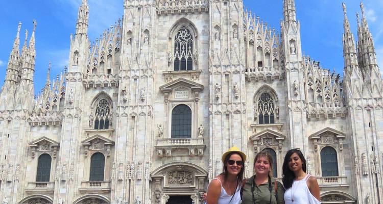 Tres viajeros sonrientes posan frente a la fachada gótica ornamentada del Duomo de Milán en un día soleado.