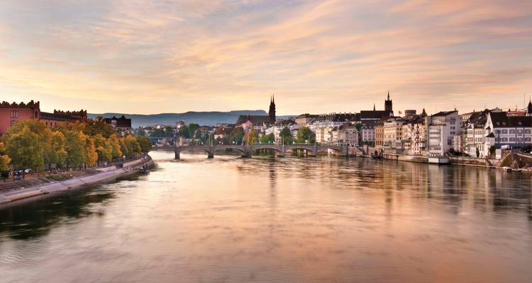 Sanfter Pastellsonnenuntergang über Basels Rheinuferpromenade mit historischer Brücke und Skyline, die sich im Wasser spiegelt.
