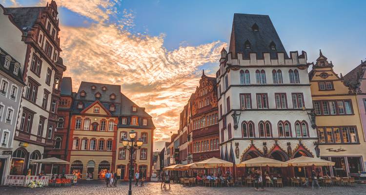 Belebter mittelalterlicher Platz in Trier zur goldenen Stunde mit verzierten Fassaden, Cafés und dramatischem wolkenbeleuchteten Himmel.