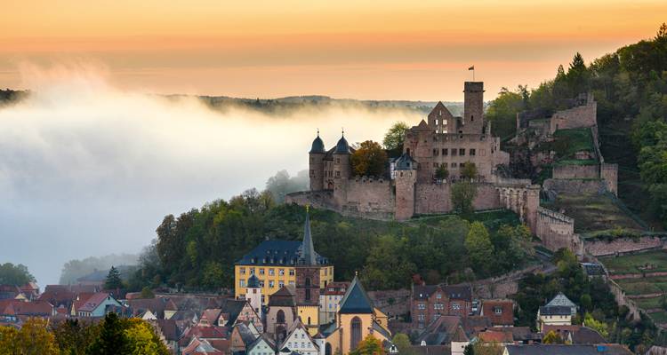Nebliger Sonnenaufgang über den Berggipfelruinen der Burg Wertheim, die sich über den Dächern der Stadt erhebt.