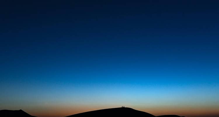 Silhouette de deux personnes assises sur une dune sous un ciel dégagé au crépuscule.