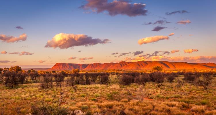 Uitgestrekte Centraal-Australische outback-vlaktes en verre tafelbergen gebaad in gouden avondlicht.