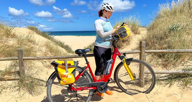 Ciclista de pie con una bicicleta eléctrica entre dunas de arena dorada y cercas de madera cerca de un mar turquesa.